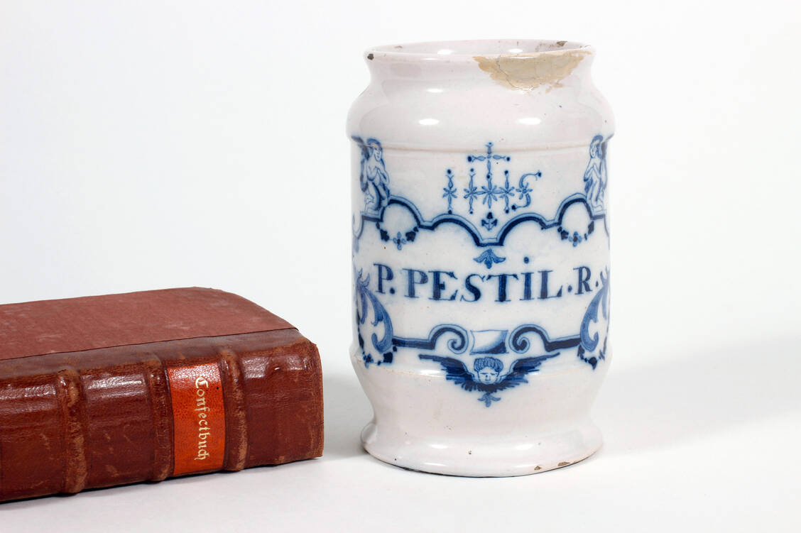 Abbildung 1: Fayence-Gefäß für »P(illulae) Pest(ilentiales) R(uffii)« aus der Residenz-Apotheke in Büren. Delft (Niederlande), um 1750 (Inv.-Nr. II E 370). Dabei das »Confect Buch unnd Hauß Apoteck« des Hermann Walter Ryff 1593, der diese Pillen ebenfalls beschreibt. (Bibl.-Sign. Ryf 1/1) / Foto: Deutsches Apotheken-Museum