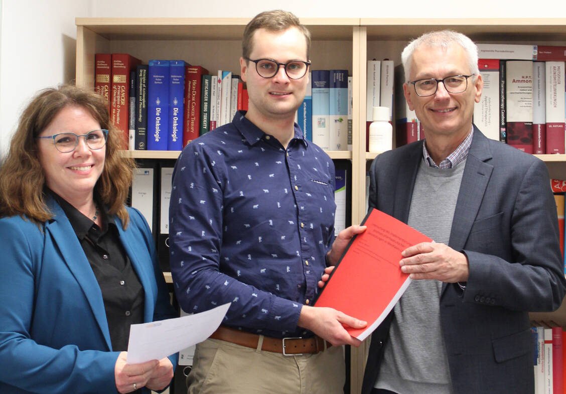 Apotheker Simon Siuts (Mitte) übergibt die erste Masterarbeit im Masterstudiengang AMTS an den Studiengangsleiter Professor Dr. Ulrich Jaehde und die Studiengangskoordinatorin Dr. Judith Hildebrand. / © privat